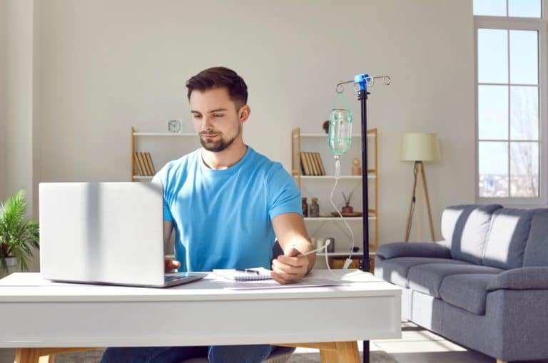 Young Man Sitting And Working On A Laptop While Having At-home IV Therapy